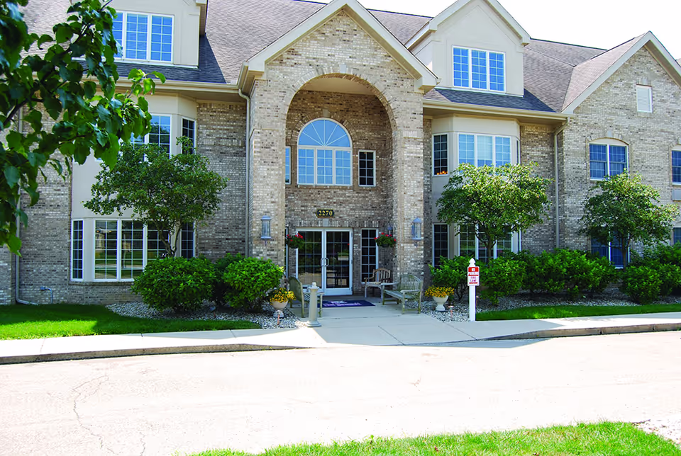 Front exterior view of a two-story brick building with large windows, a central arched entrance with glass double doors, surrounded by green bushes and trees, and a paved driveway in front.