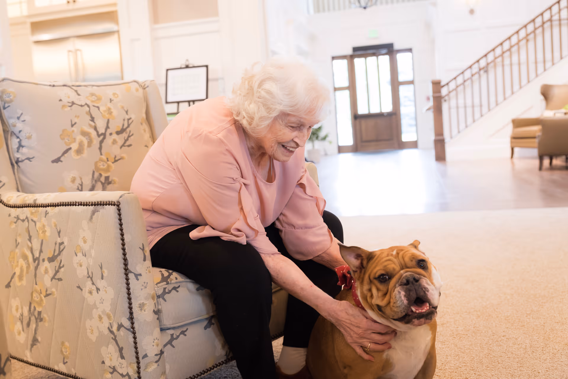 An elderly woman with white hair wearing a pink blouse and black pants is sitting on a floral patterned armchair, smiling and petting a brown and white bulldog in a spacious, well-lit living room with a staircase and large wooden door in the background.