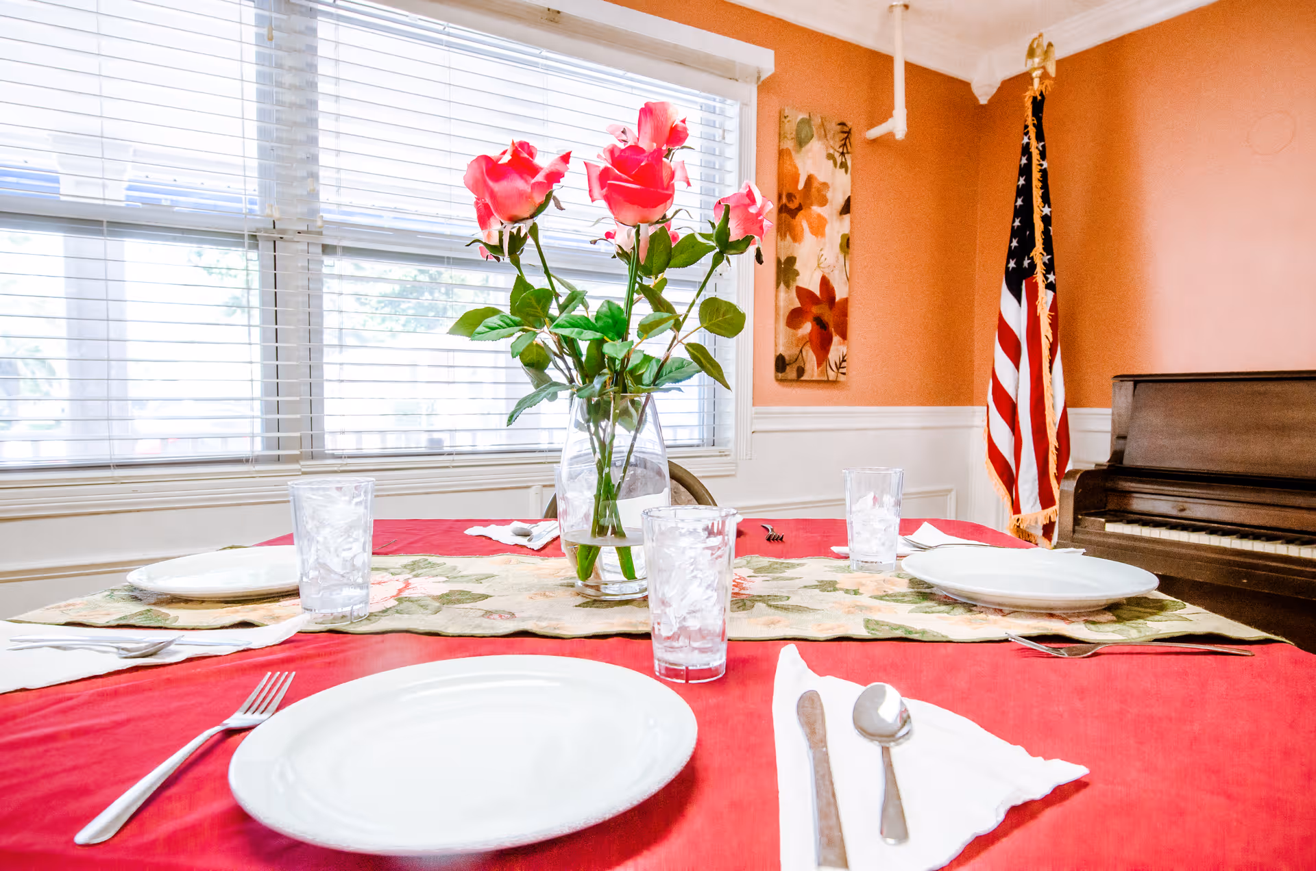 A dining table set with white plates, silverware, and glasses of ice water on a red tablecloth with a floral runner. A vase with pink roses is in the center of the table. In the background, there is a window with blinds, an American flag, a piano, and a floral wall decoration on a peach-colored wall.