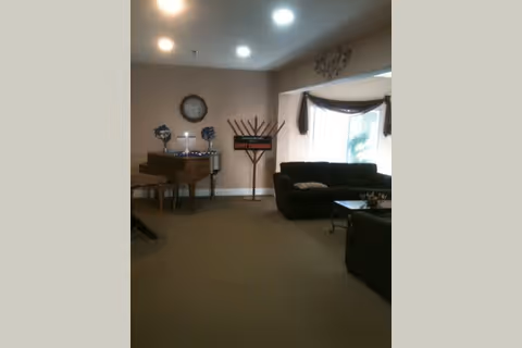 A cozy living room area with a dark sofa and armchair, a coffee table, a piano with decorative items on top, a large clock on the wall, and a menorah with a 'Happy Chanukah' sign. The room has beige walls and carpet, recessed ceiling lights, and a large window with curtains letting in natural light.