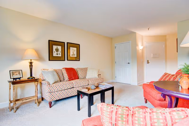 A cozy living room with a patterned sofa and two red armchairs. There is a wooden side table with a lamp and a framed photo on it, and a small black coffee table with books and decorative items. The walls are painted light beige with two framed botanical prints hanging above the sofa. The room has a carpeted floor and a white door in the background.