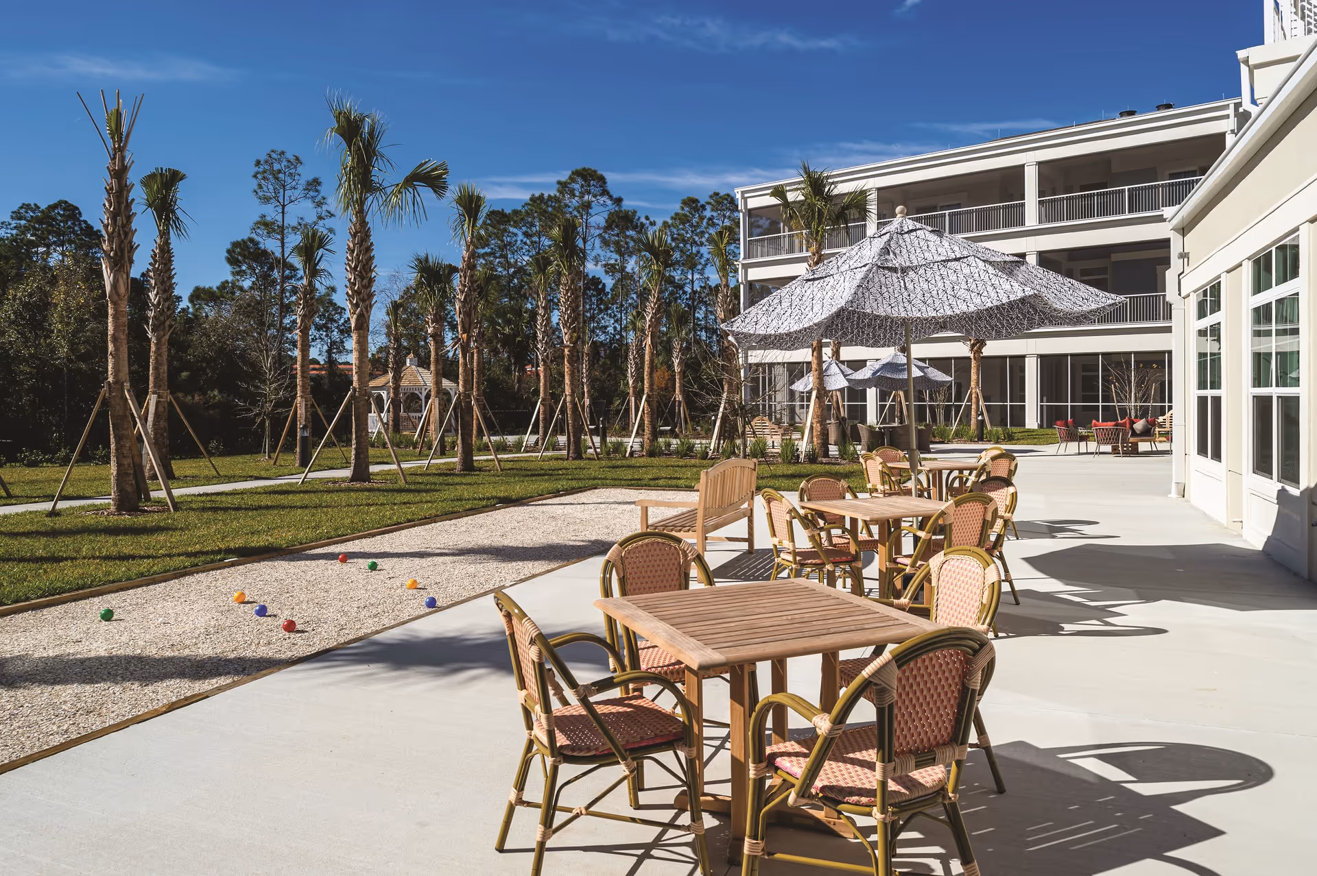 Outdoor patio area at Sancerre at Palm Coast with wooden tables and chairs under large patterned umbrellas. There are palm trees planted in the grassy area next to a bocce ball court with colorful balls. A multi-story building with balconies is visible in the background under a clear blue sky.