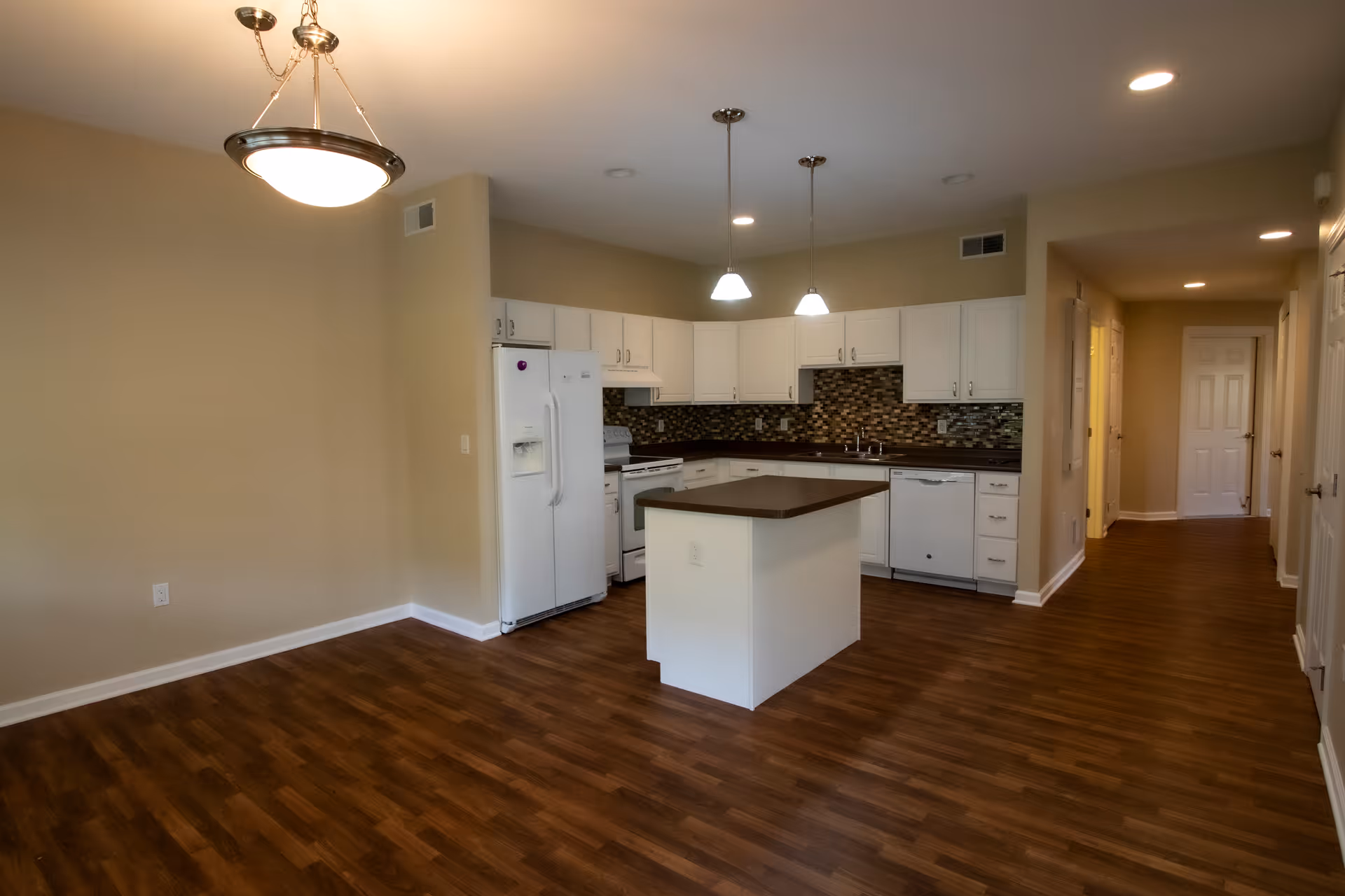 A modern kitchen interior featuring white cabinetry, a central island, and a tiled backsplash. The kitchen includes a refrigerator, stove, and dishwasher, with warm wood flooring and pendant lighting.