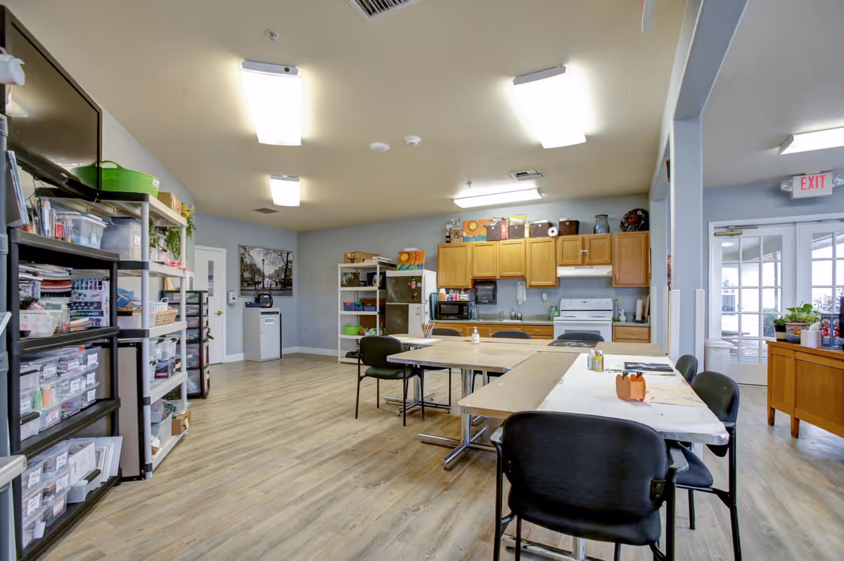 A spacious room with light wood flooring and fluorescent ceiling lights, featuring a kitchenette with wooden cabinets, a white stove, microwave, and refrigerator. Several tables and black chairs are arranged in the center, with shelves on the left holding various craft supplies and materials. A door with glass panels leads to an outdoor area, and an exit sign is visible above it.