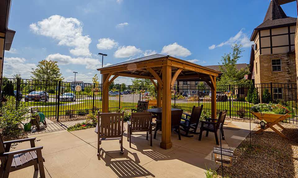 Outdoor patio area at a senior living facility with a wooden pergola covering a round table and several chairs. The area is surrounded by a black metal fence, with a parking lot and trees visible in the background under a partly cloudy blue sky.