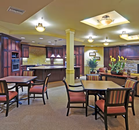 Interior view of a senior living facility dining area with multiple wooden tables and chairs with red cushions. The room features a kitchen area with dark wood cabinetry, a tiled backsplash, and pendant lighting. There is a decorative stone wall with yellow flowers and plants on a side table, and ceiling lights provide warm illumination.