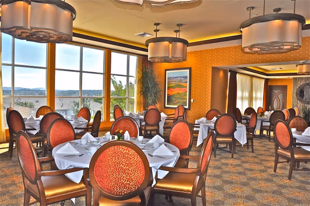 A bright dining room with multiple round tables covered in white tablecloths, each set with folded napkins and silverware. The chairs have red patterned upholstery and wooden frames. Large windows let in natural light and offer a view of the outdoors. The room features warm yellow and orange walls, modern ceiling lights, a framed landscape painting, and decorative plants.