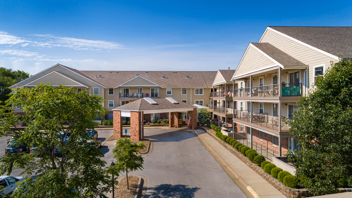 Exterior view of Twin Oaks Assisted Living facility showing a multi-story building with balconies, a covered entrance supported by brick pillars, a driveway, parked cars, and surrounding greenery under a clear blue sky.