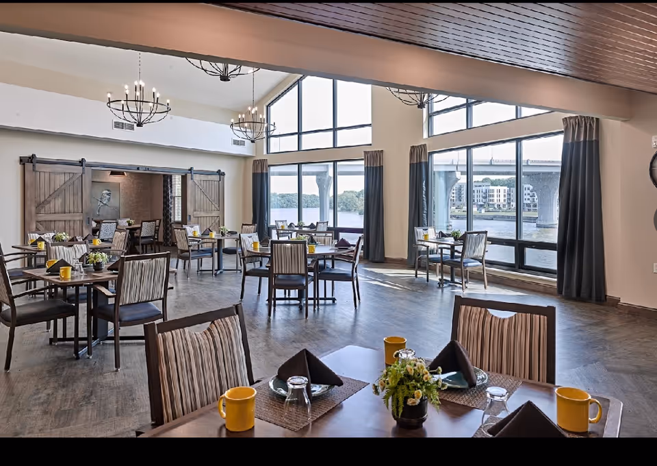Bright dining room with multiple set tables and large floor-to-ceiling windows overlooking a river and bridge.
