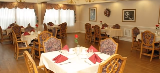 A dining room with multiple wooden tables and chairs, each table covered with a white tablecloth and set with red folded napkins, glasses, and a single red rose in a vase. The room has large windows with sheer curtains, framed artwork on the walls, and warm lighting from chandeliers.