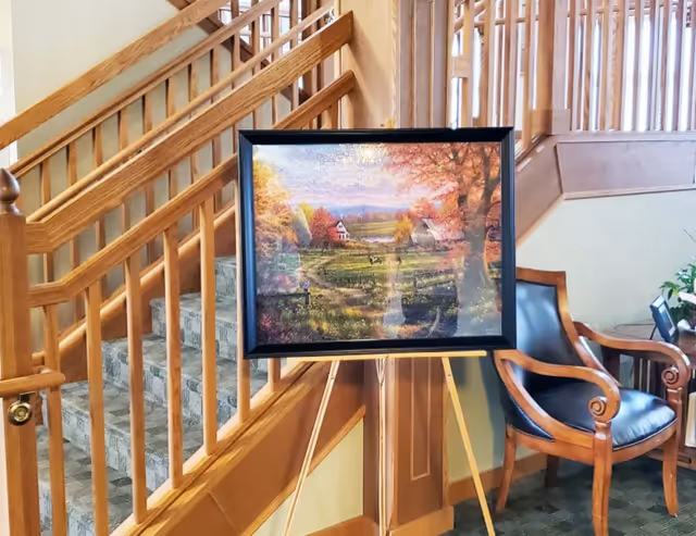 A framed landscape painting displayed on an easel in a lobby area beside a wooden staircase and a leather chair.
