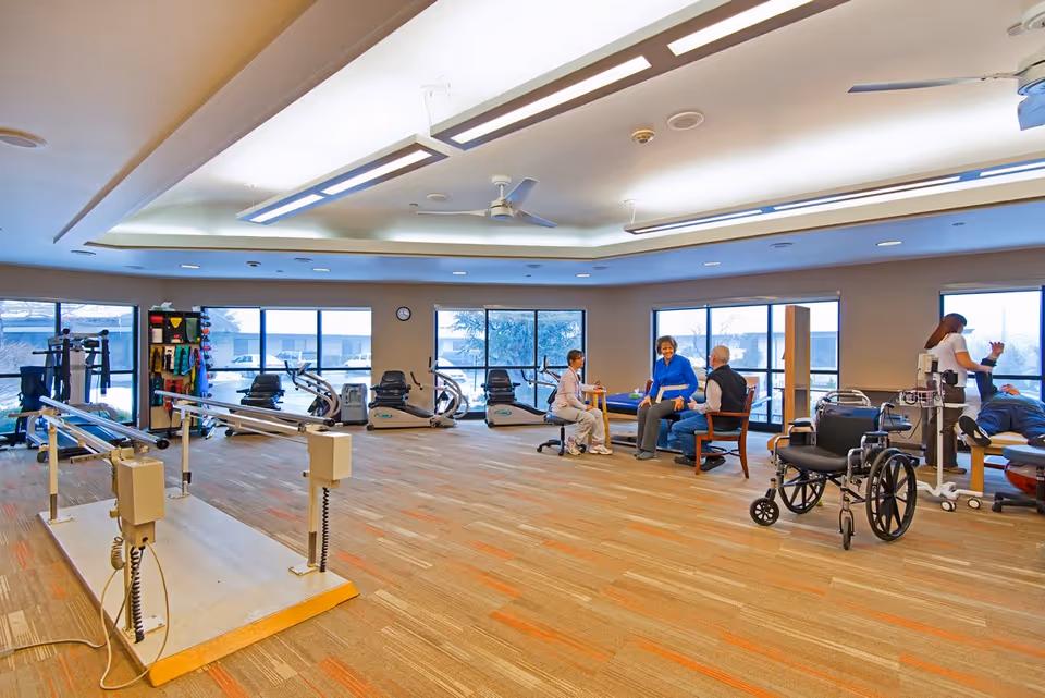 A spacious rehabilitation or physical therapy room with exercise equipment including parallel bars and stationary bikes. Several people are engaged in therapy activities, with one person assisting a patient on a therapy table and two others seated and conversing. The room has large windows letting in natural light and a wheelchair is visible in the foreground.