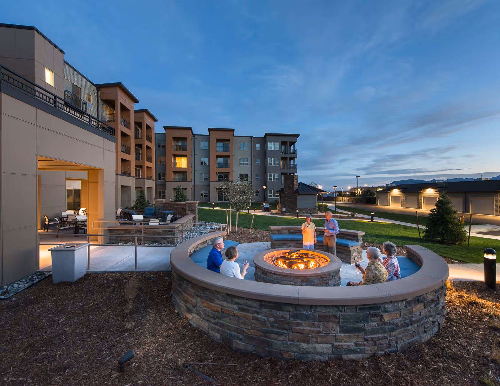 Outdoor evening scene at Revel Province senior living facility showing a group of elderly people gathered around a circular stone fire pit. The background features a multi-story residential building with balconies and outdoor seating areas under a twilight sky.
