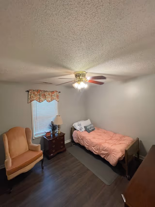 A small bedroom with a single bed covered in a pink comforter and a pillow with the text 'Better together'. There is a wooden nightstand with a lamp and decorative items next to the bed. A peach-colored upholstered armchair is placed near a window with floral valance curtains. The room has dark wood flooring and a ceiling fan with lights.