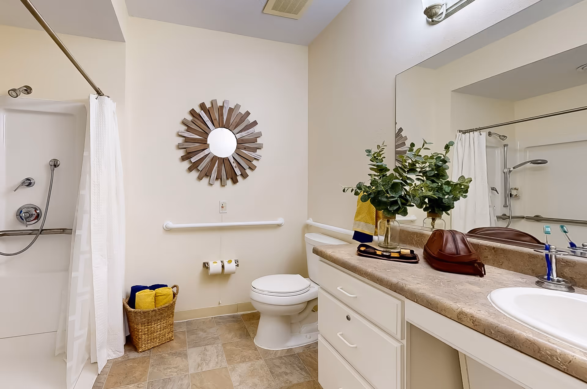 A clean and well-lit bathroom featuring a shower with a white curtain, a toilet, a countertop with a sink, and a large mirror. There is a decorative round mirror on the wall above the toilet, a basket with rolled towels on the floor, and a vase with green foliage on the countertop.