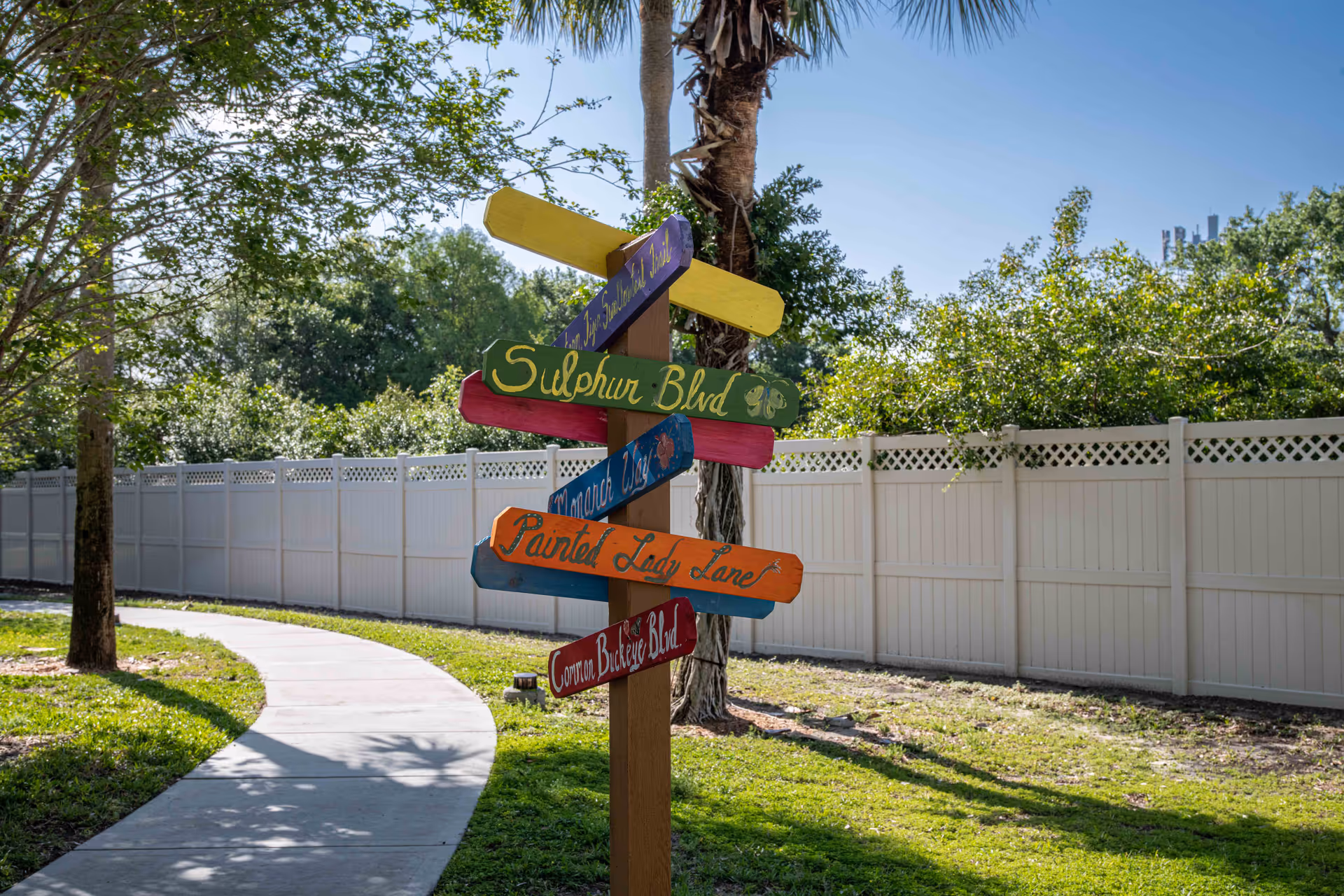 A colorful wooden signpost with multiple directional signs displaying names such as Sulphur Blvd, Painted Lady Lane, and Common Buckeye Blvd, situated next to a curved concrete pathway in a green outdoor area with trees and a white fence in the background under a clear blue sky.
