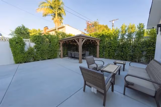 Outdoor patio area with concrete flooring, featuring a wooden pergola with string lights and a dining table with chairs underneath. In the foreground, there is a seating area with cushioned chairs and a small table. The space is surrounded by tall green hedges and a white fence, with a palm tree and other trees visible in the background.