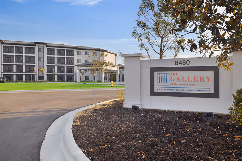 Entrance sign for The Gallery at Hacienda Lakes by a driveway with the multi-story senior living building in the background.