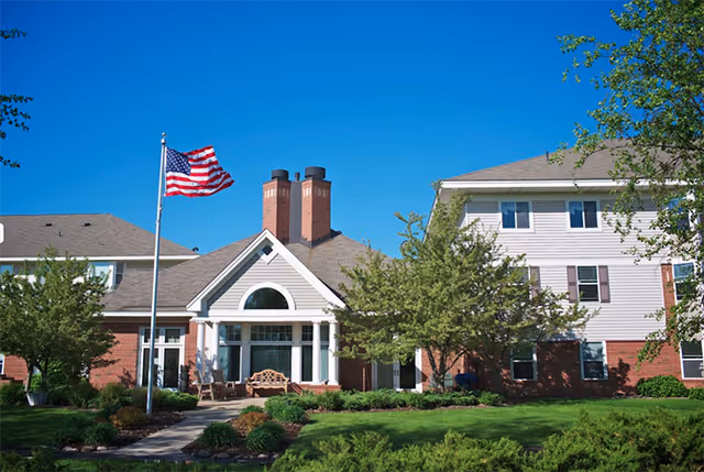 Exterior view of Oak Meadows Senior Living facility showing a well-maintained lawn, trees, and shrubs. The building features a combination of brick and light-colored siding with multiple windows and a prominent entrance with white columns. An American flag is flying on a flagpole near the entrance under a clear blue sky.
