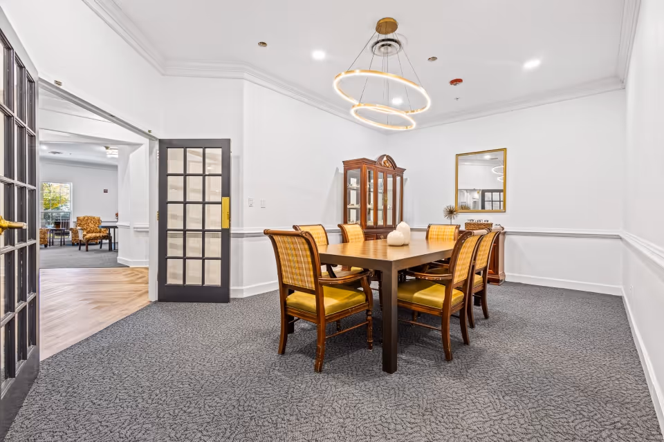 A well-lit dining room in a senior living facility featuring a rectangular wooden dining table surrounded by six chairs with yellow cushions and plaid backs. A modern circular chandelier hangs above the table. Against the wall is a wooden china cabinet with glass doors and a mirror above a small sideboard. The room has white walls, gray carpet, and an open doorway leading to another room with seating and natural light.