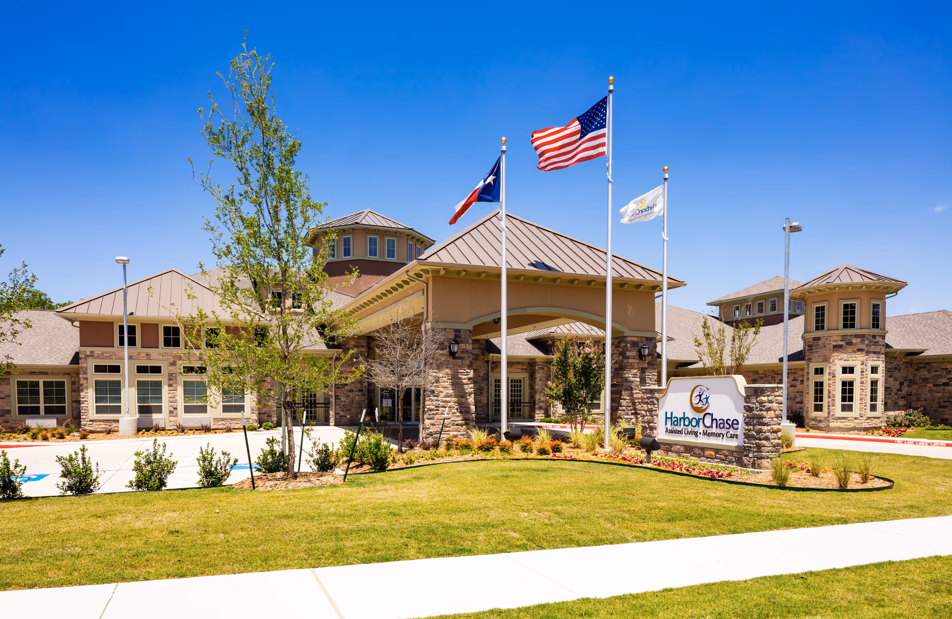 Exterior view of a senior living facility building with a well-maintained lawn and landscaping. The building features stone and beige siding with multiple windows and a covered entrance. Three flagpoles stand in front, displaying the American flag, the Texas state flag, and a facility flag. A sign near the entrance reads 'HarborChase Assisted Living Memory Care.'