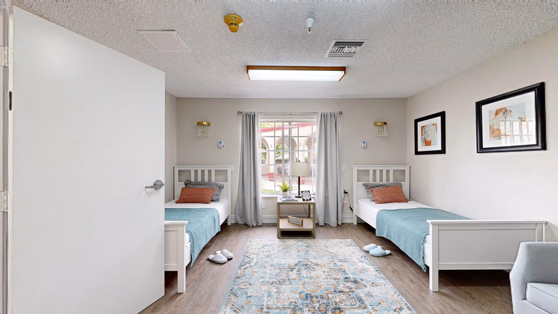 A bright and clean bedroom with two single beds, each with a teal blanket and orange pillow. Between the beds is a small table with a lamp, plant, and a welcome sign. A window with light gray curtains looks out to an outdoor patio area. The room has light-colored walls, wooden flooring, a patterned area rug, and two framed abstract artworks on the right wall.