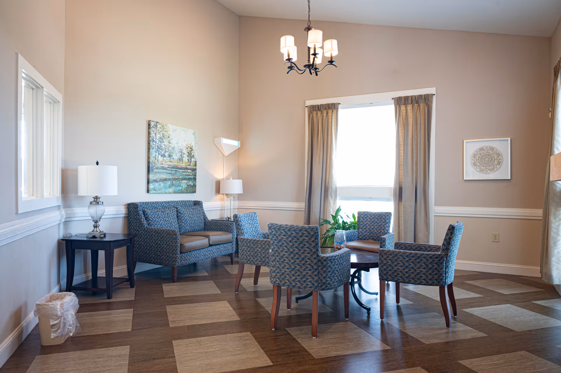 A bright and cozy sitting area in a senior living facility with a small sofa, four patterned armchairs arranged around a round wooden table, a side table with a lamp, a floor lamp, framed artwork on the walls, and a large window with curtains letting in natural light.