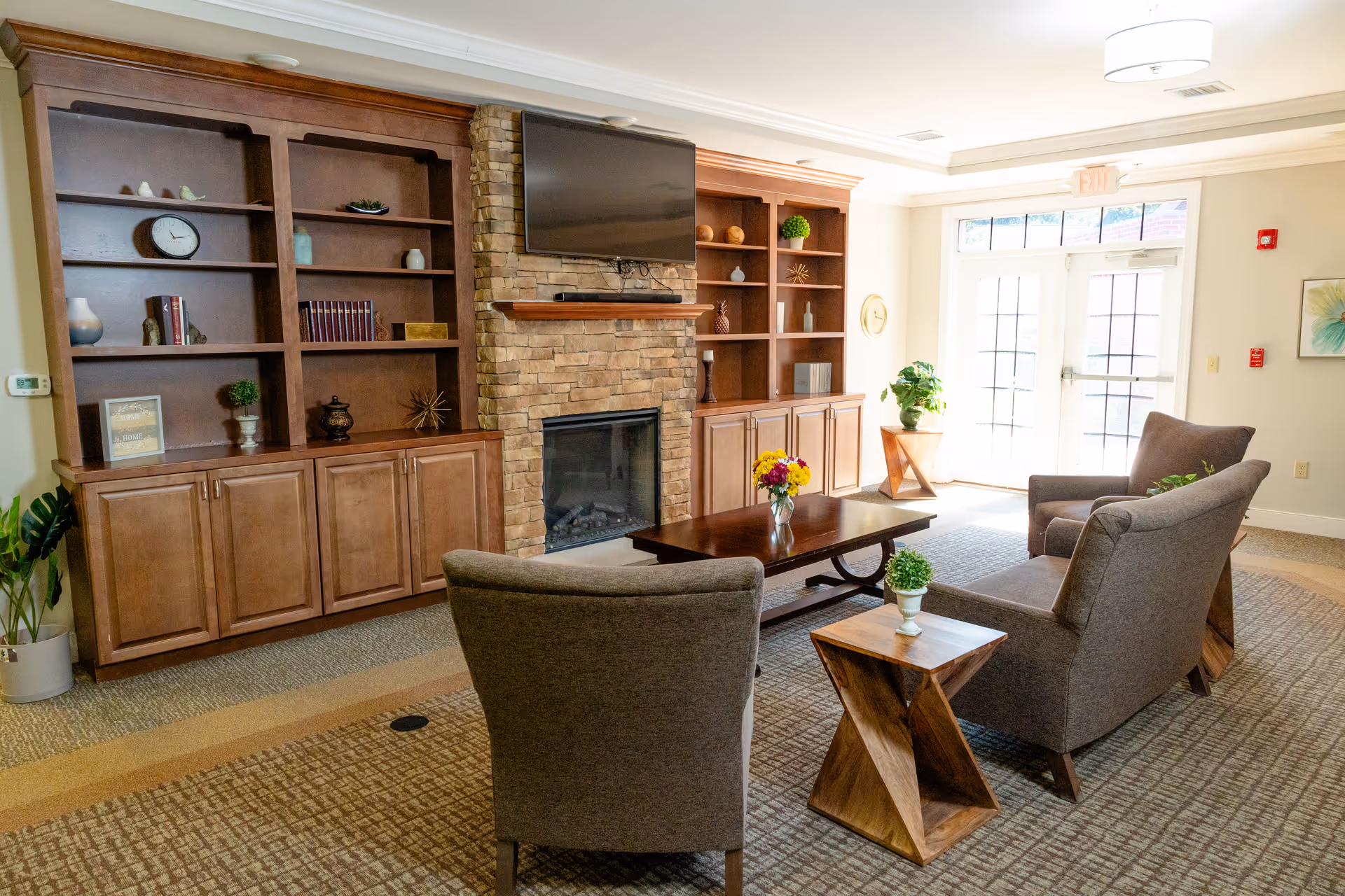 A cozy senior living common area with two gray armchairs and a matching loveseat arranged around a wooden coffee table with a vase of flowers. Behind the seating area is a stone fireplace with a mounted flat-screen TV above it, flanked by built-in wooden shelves and cabinets. The room is well-lit with natural light coming through glass doors and has a carpeted floor with a modern pattern.