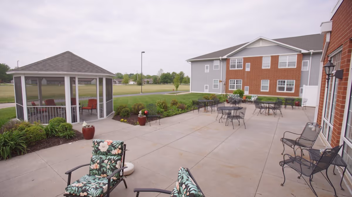 Outdoor patio area at John M. Evans Supportive Living Community featuring multiple metal tables and chairs, cushioned chairs with floral patterns, a screened gazebo with red cushioned chairs inside, and landscaped greenery surrounding the concrete patio. The building exterior is visible with brick and gray siding.