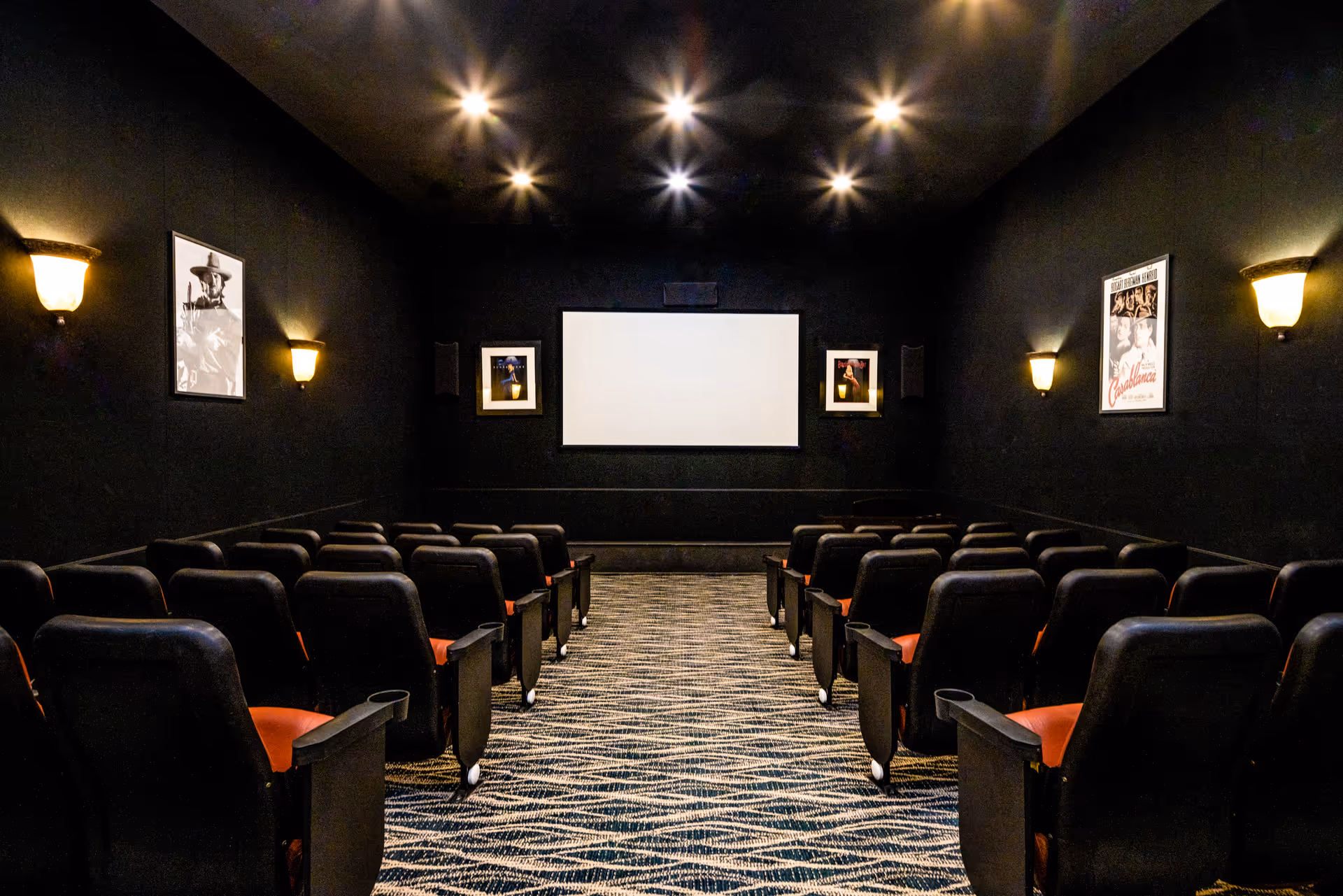 Interior view of a small movie theater with rows of black and red cushioned seats facing a large blank white screen. The walls are black with framed movie posters and wall-mounted lights providing warm illumination. The ceiling has several recessed lights.
