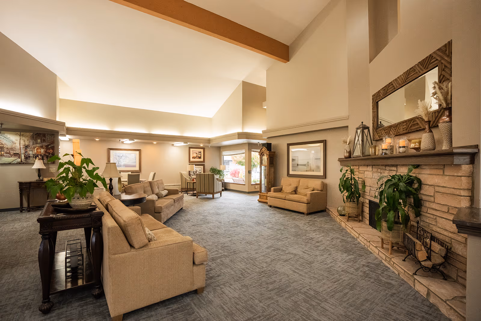 A spacious and well-lit living room area with beige sofas, a stone fireplace decorated with plants, candles, and vases, a large mirror above the fireplace, framed artwork on the walls, and a grandfather clock near a window showing outdoor foliage.