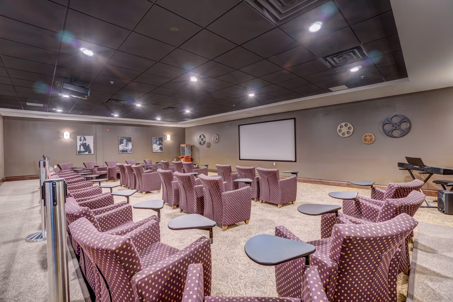 A theater-style room with multiple rows of purple armchairs with small attached tables, facing a large blank projection screen on the wall. The room has a dark ceiling with recessed lighting, decorative film reels on the wall, black and white portraits, a popcorn machine in the back corner, and a keyboard on the right side.