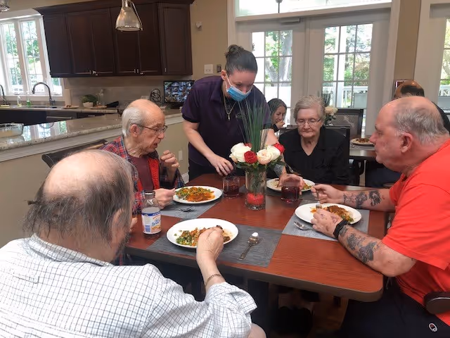 Four residents eat a meal around a dining table while a masked staff member serves them in a communal dining room.