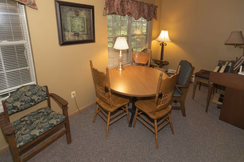 A cozy interior room with a round wooden table surrounded by four wooden chairs. There is a lamp on the table and another floor lamp in the corner. A window with blinds and a decorative valance is on the wall behind the table. To the left, there is a cushioned wooden armchair with floral upholstery. On the right side, there is a piano with sheet music on it. The walls are painted beige and the floor is carpeted.