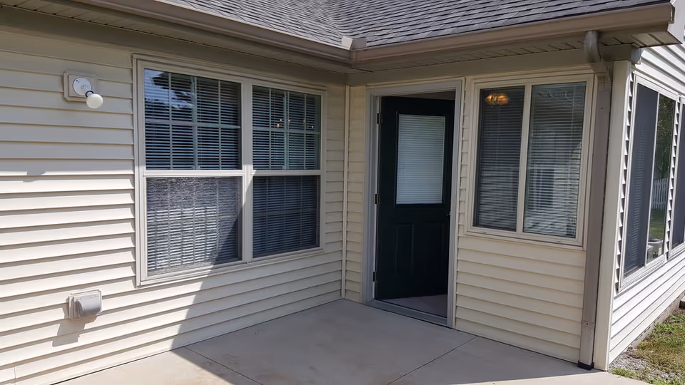 Exterior of a beige vinyl-sided unit showing windows, a dark green door and a small concrete patio.