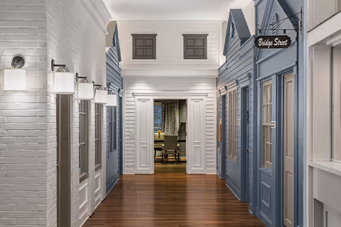 Indoor hallway designed to resemble a small village street with house-like facades painted in white and blue, wooden floors, wall-mounted lights, and a sign reading 'Bridge Street'. At the end of the hallway, there is an open doorway leading to a room with a table and chairs.