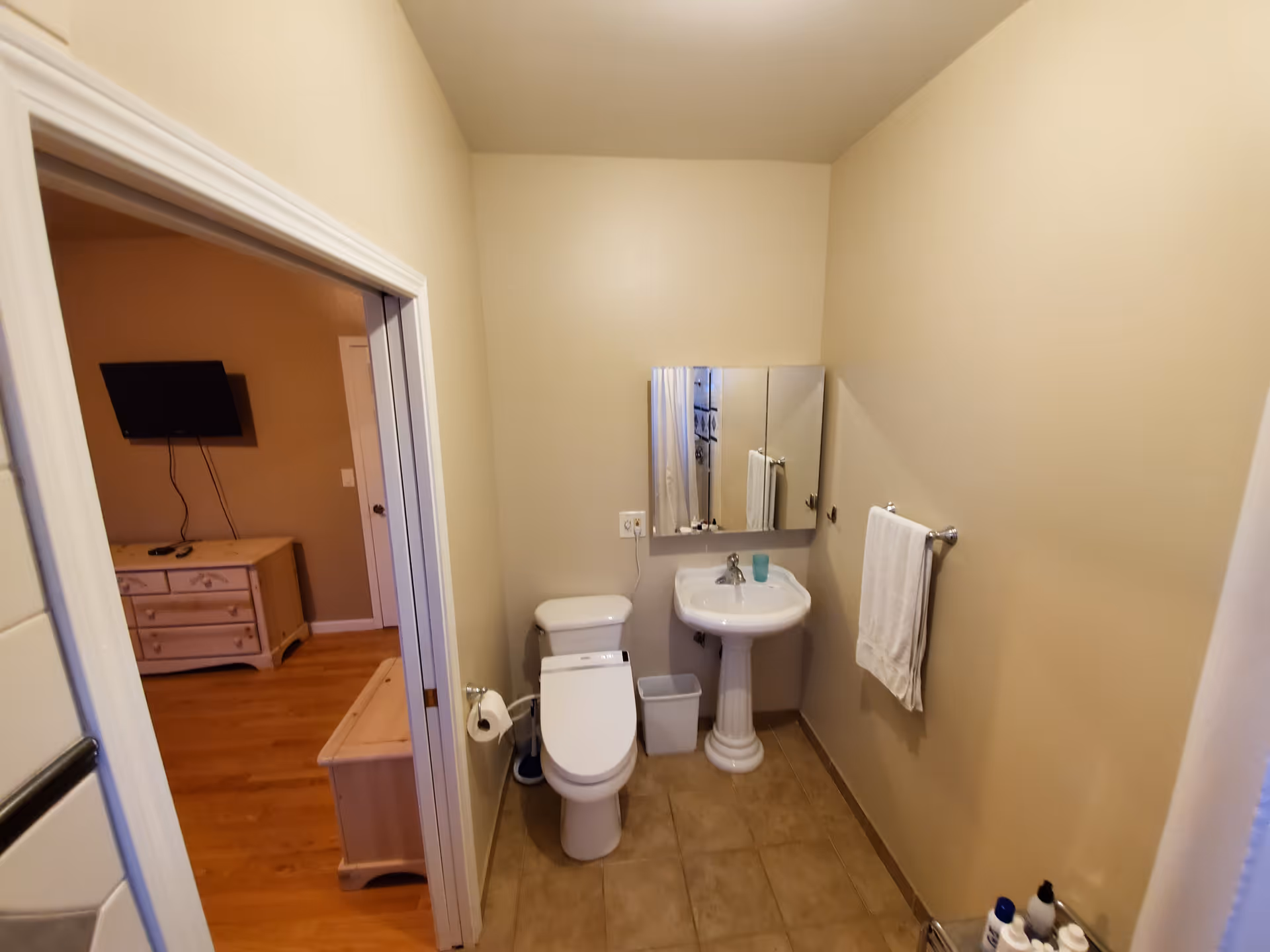 A small bathroom with beige walls and tiled floor featuring a white toilet, a pedestal sink with a mirror cabinet above it, a towel rack with a white towel, and a small trash bin. The bathroom opens into a room with wooden flooring, a dresser, and a wall-mounted TV.