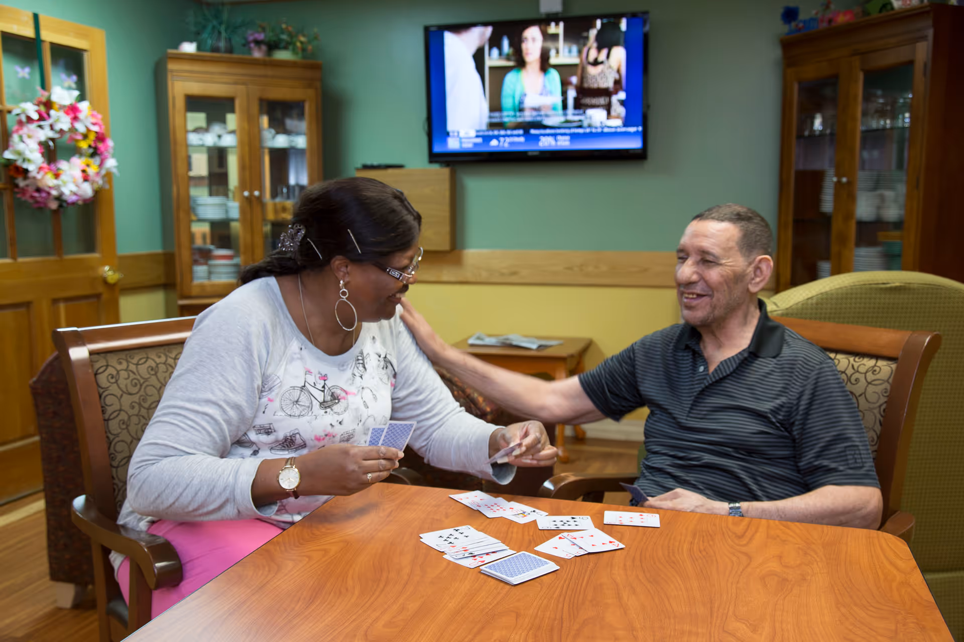Two people sitting at a wooden table playing cards in a cozy room with wooden furniture and a television mounted on the wall in the background. One person is smiling and reaching out to touch the other's shoulder.