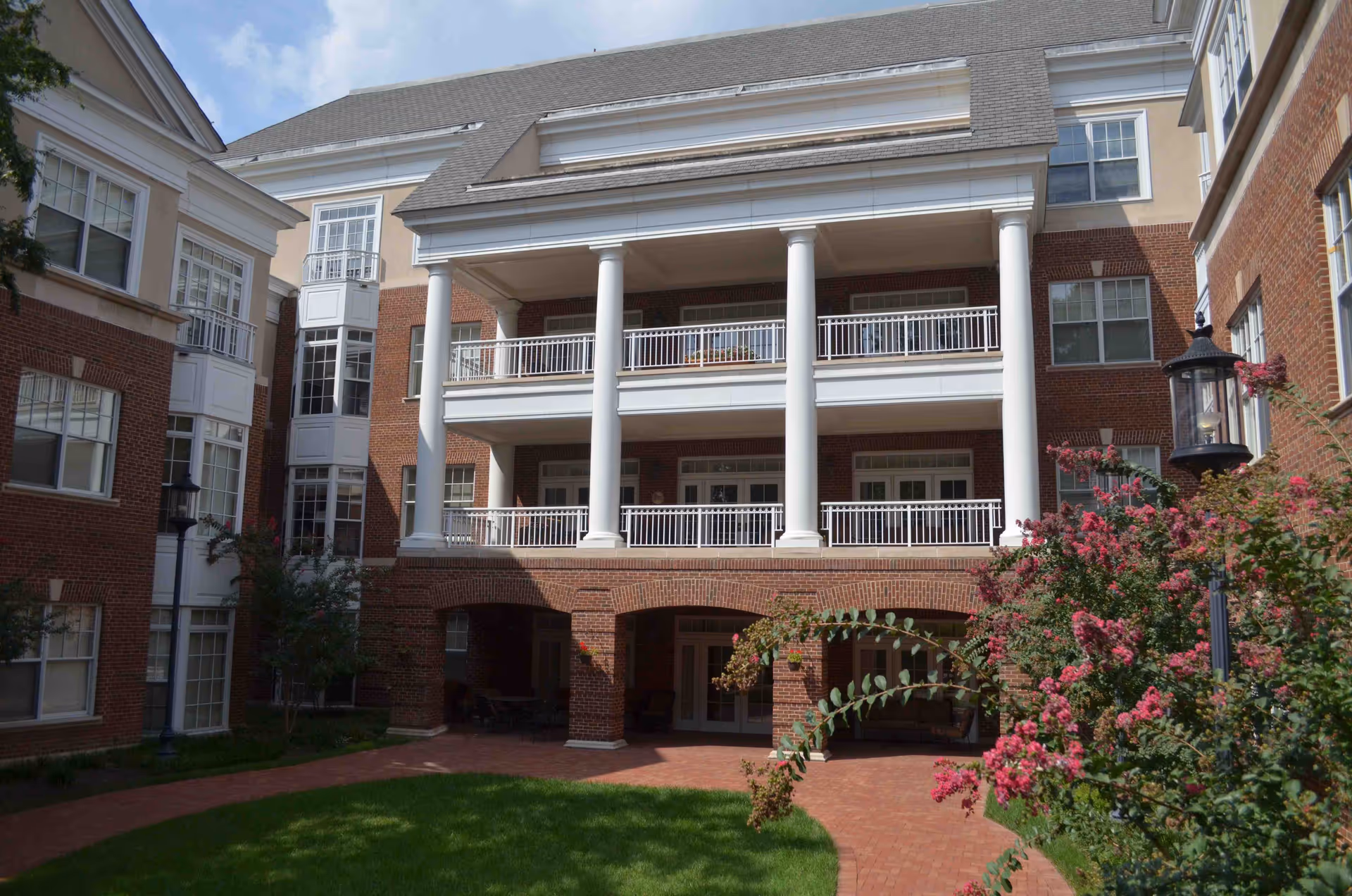 Exterior view of a multi-story assisted living facility with red brick walls, white columns, balconies, and large windows. There is a well-maintained green lawn and flowering bushes in the foreground under a partly cloudy sky.