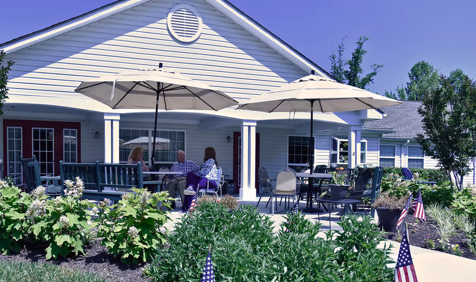 Outdoor patio area at Great Falls Memory Care by Artis with tables, chairs, and large umbrellas. Three people are seated and conversing under the umbrellas. The patio is surrounded by green plants and small American flags, with a white building in the background.