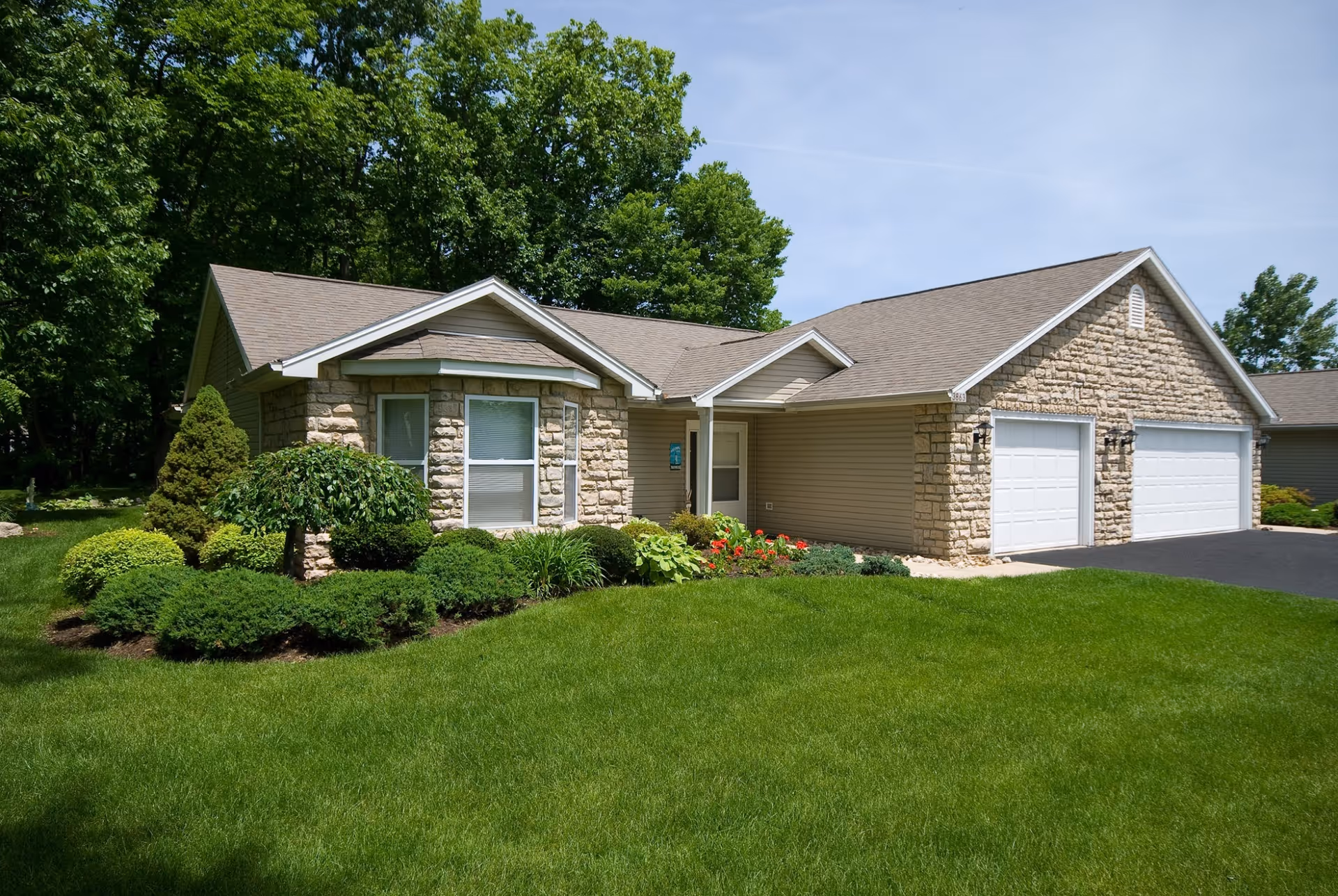 Single-story stone-and-siding house with an attached three-car garage, manicured lawn and shrubs under a clear sky.