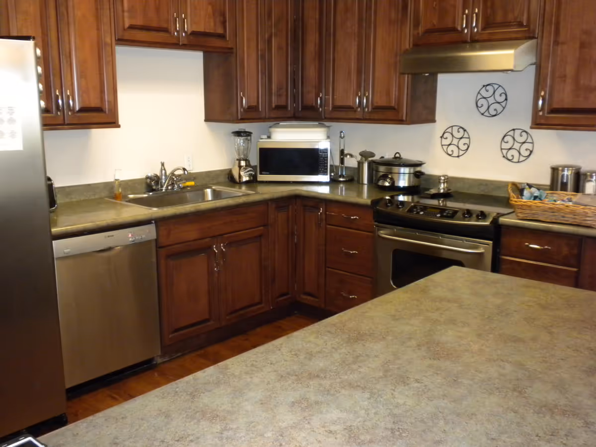 A kitchen with wooden cabinets, a stainless steel refrigerator, dishwasher, stove, and microwave. The countertop has a blender, a slow cooker, and a basket with various items. Decorative metal wall hangings are on the wall above the stove.