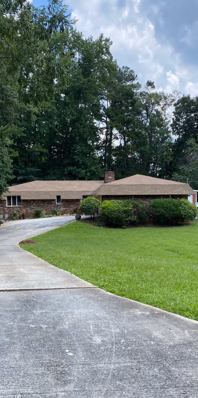 A single-story brick residential building with a brown shingled roof surrounded by green grass and bushes. A curved concrete driveway leads up to the house, and tall trees are visible in the background under a partly cloudy sky.