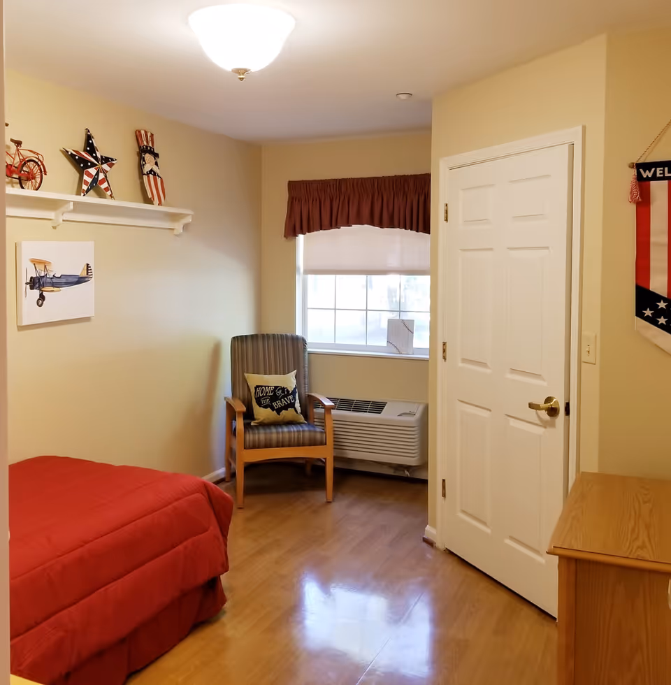 A small bedroom with a single bed covered in a red comforter, a wooden chair with a striped cushion and a pillow that says 'Home of the Brave', a window with a brown valance and a white blind, a white door with a gold handle, and a wooden dresser. The walls are beige and there are patriotic decorations including a red bicycle, a star, and a figure on a shelf, as well as a painting of a blue airplane on the wall.