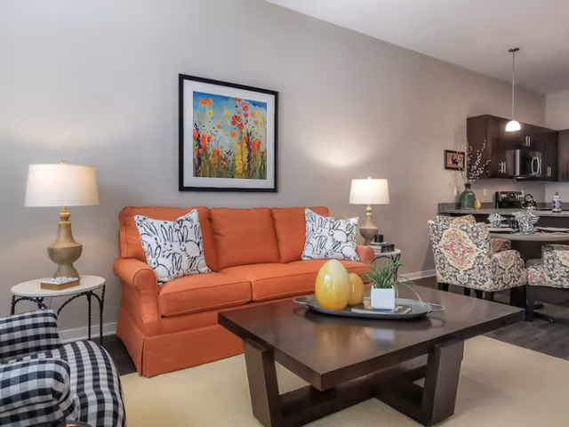 A cozy living room with an orange sofa adorned with two white pillows featuring black bunny designs. There is a dark wooden coffee table with decorative items including a small plant and two yellow vases. To the left, there is a black and white checkered armchair and a small round side table with a lamp. The background shows a kitchen area with dark cabinets, a microwave, and a countertop with floral-patterned chairs. A colorful floral painting hangs on the wall above the sofa.