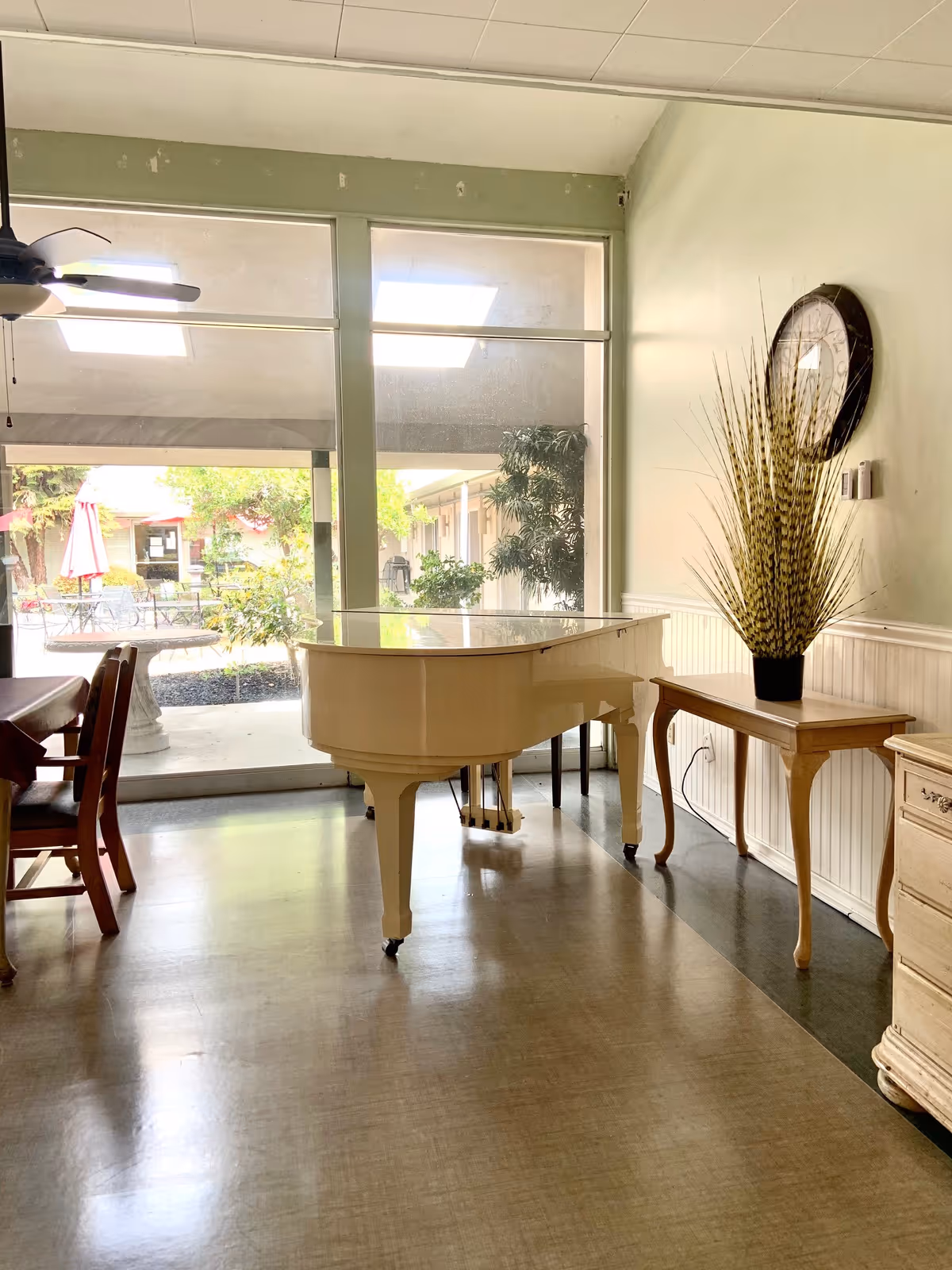 Interior room with a white grand piano in the center, a wooden table with a tall decorative plant on the right, wooden chairs and a table on the left, large windows showing an outdoor garden area with trees and umbrellas, and a ceiling fan above.