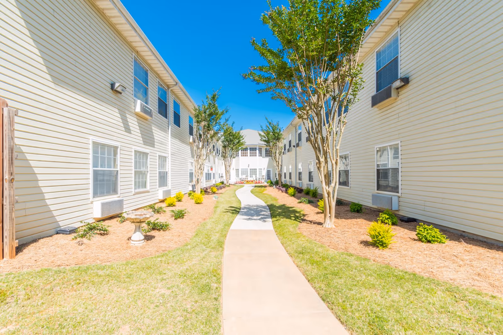 A bright outdoor walkway between two beige two-story buildings with multiple windows and air conditioning units. The path is flanked by small landscaped areas with trees, shrubs, and mulch under a clear blue sky.