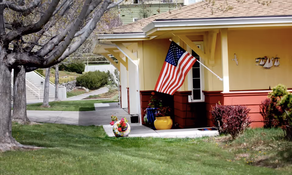 Exterior view of a building corner with a yellow and red facade, an American flag mounted on the wall, decorative flower pots on the ground, and some bushes and trees nearby on a grassy lawn.