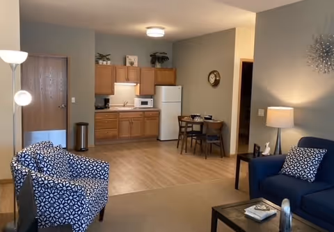 A cozy senior living apartment interior featuring a small kitchen with wooden cabinets, a white refrigerator, microwave, and a small dining table with two chairs. The living area has a blue armchair with a patterned cushion, a patterned accent chair, a floor lamp, a table lamp on a side table, and a coffee table with books. The walls are painted in neutral tones and there is a decorative clock and wall art.