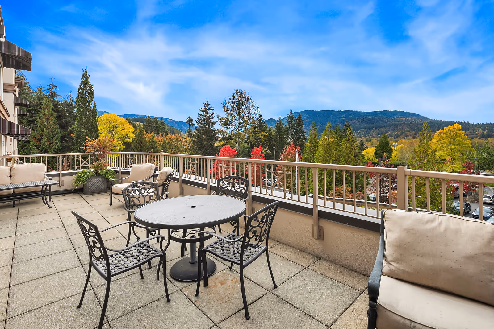 Outdoor patio area with metal table and chairs, cushioned seating, potted plants, and a railing overlooking a scenic view of trees with autumn foliage and distant mountains under a blue sky.
